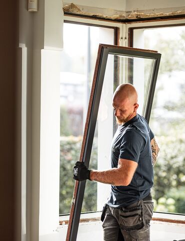 Bald man in blue shirt dismantling wooden window from residential construction site.